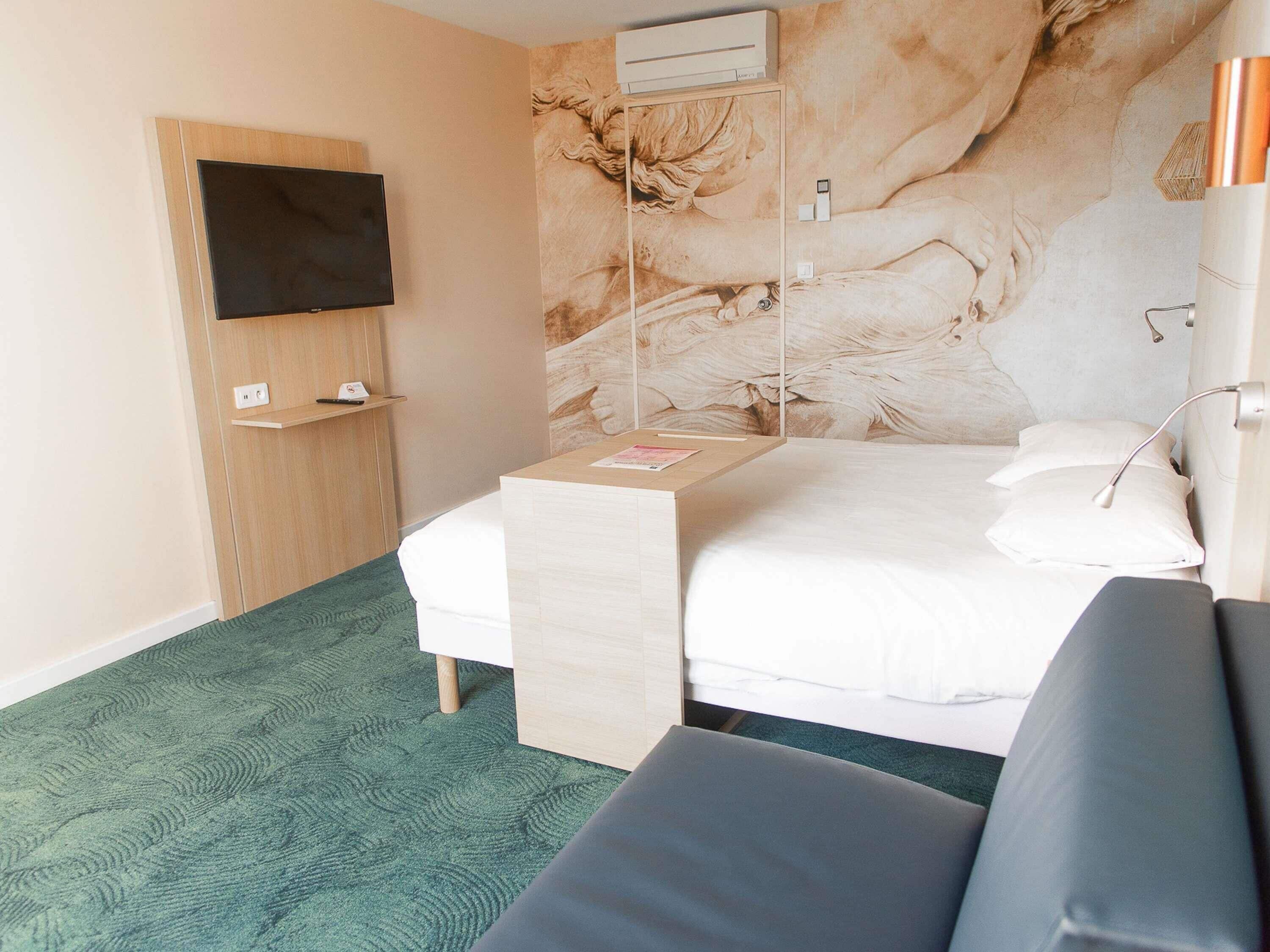 A hotel room with a bed, a TV mounted on the wall, a small table with a magazine, and a green carpet.