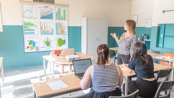 Un aula con un profesor dando clase a estudiantes, escritorios, sillas y una pizarra con gráficos.