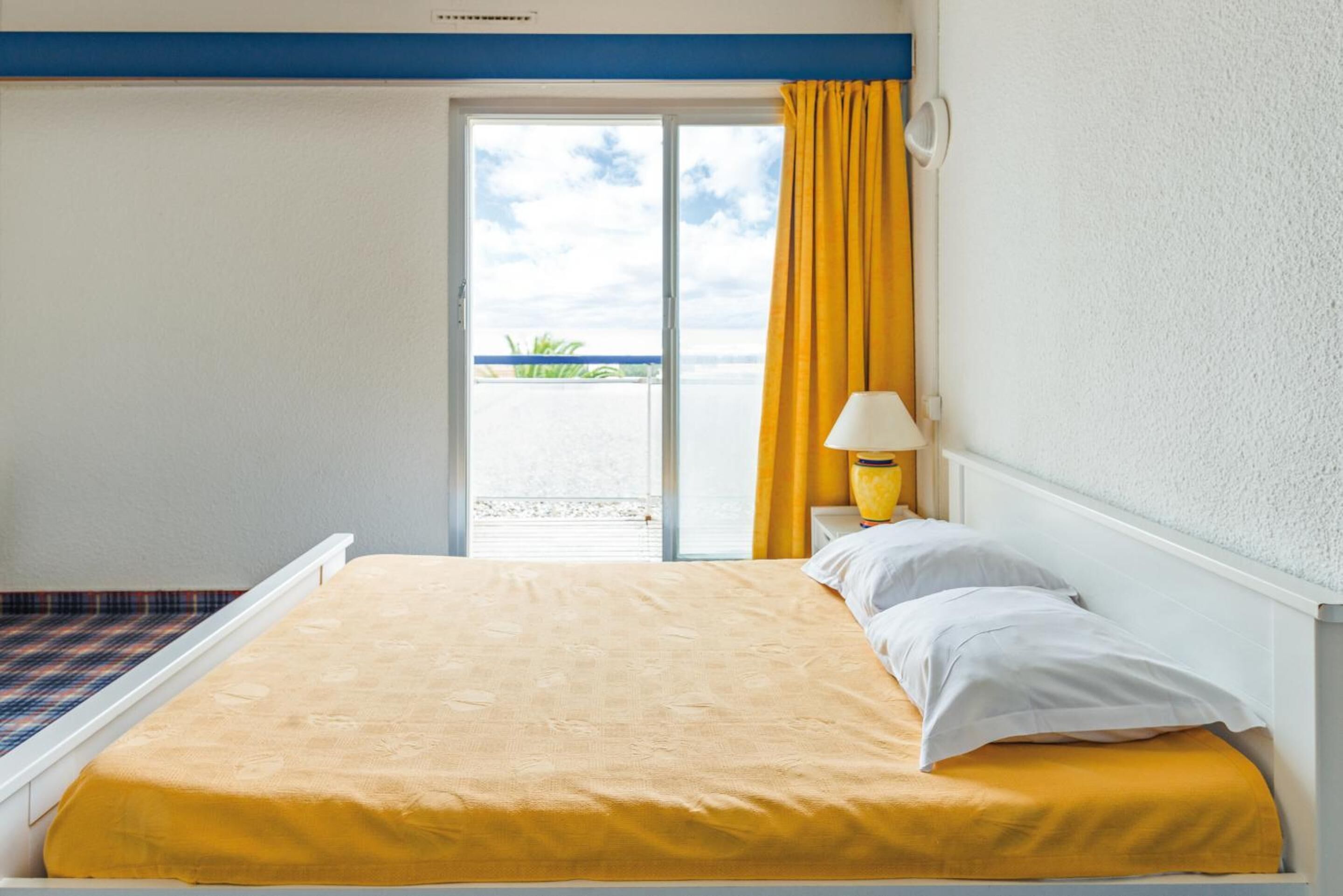 A hotel room with a bed, a nightstand with a lamp, a window with yellow curtains, and a view of the beach.