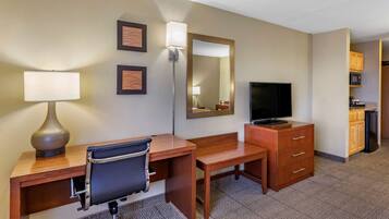 Habitación de hotel con escritorio de madera, una silla, televisor, espejo y kitchenette con microondas y armarios.