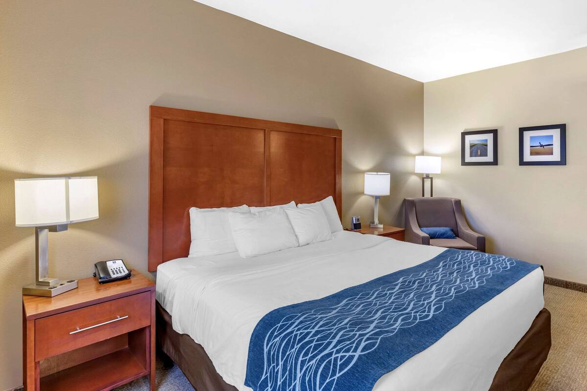 A hotel room with a large bed, a wooden headboard, a nightstand with a lamp, a chair, and two framed pictures on the wall.