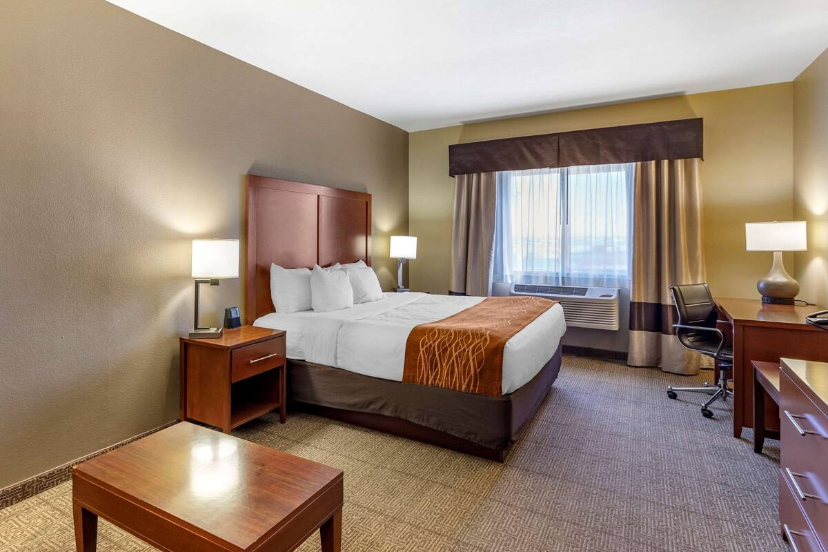 A hotel room with a large bed, two bedside tables with lamps, a desk with a chair, and a view of the city through the window.