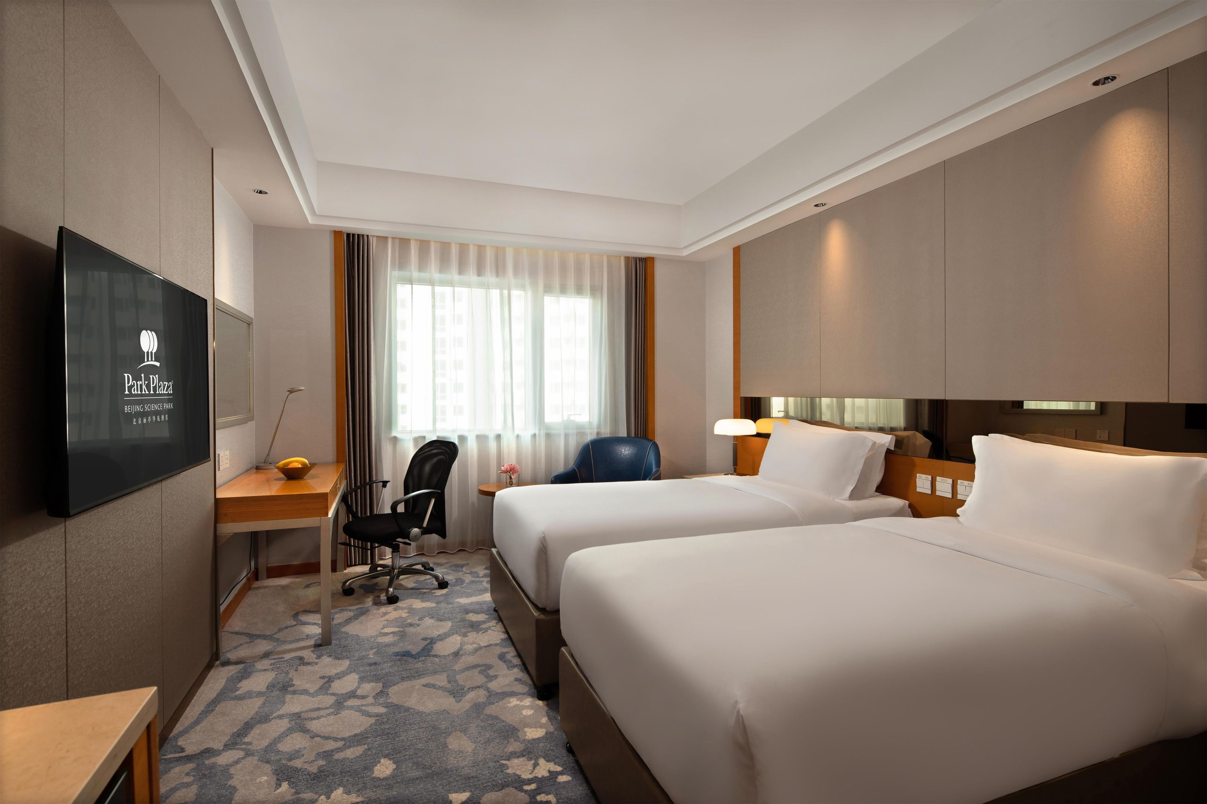 A hotel room with two beds, a desk with a chair, a TV, and a window with curtains.