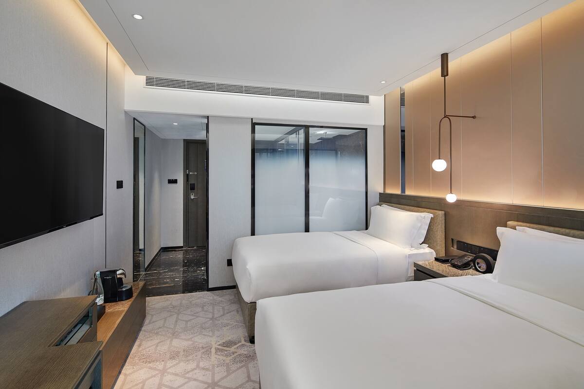 Een moderne hotelkamer met twee bedden, een flatscreen-tv, een nachtkastje en een wandlamp.