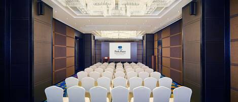 A conference room with rows of white chairs, a large chandelier, and a screen displaying 'Park Plaza'.