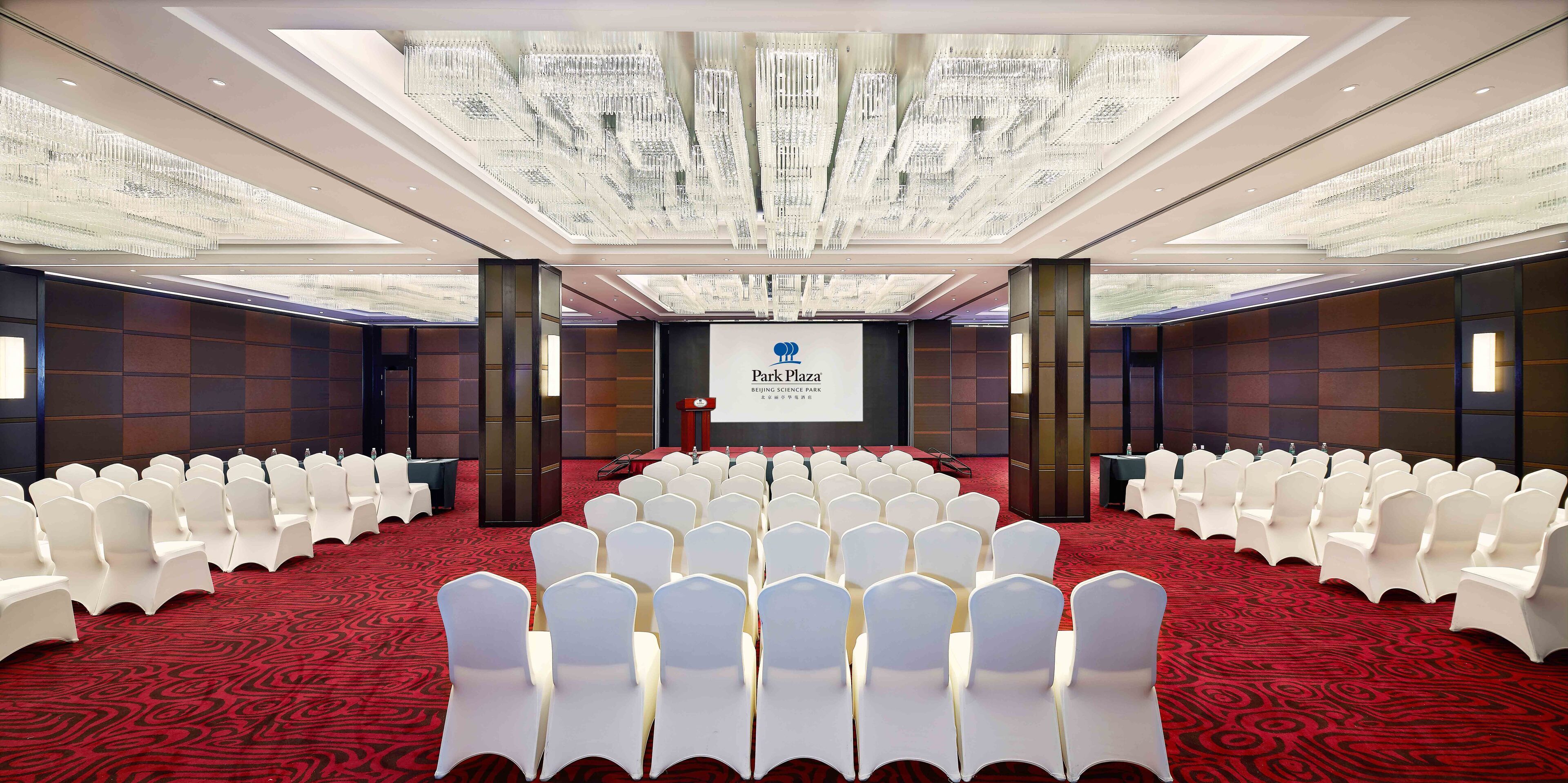 A conference room with rows of white chairs, a large screen, and a chandelier.