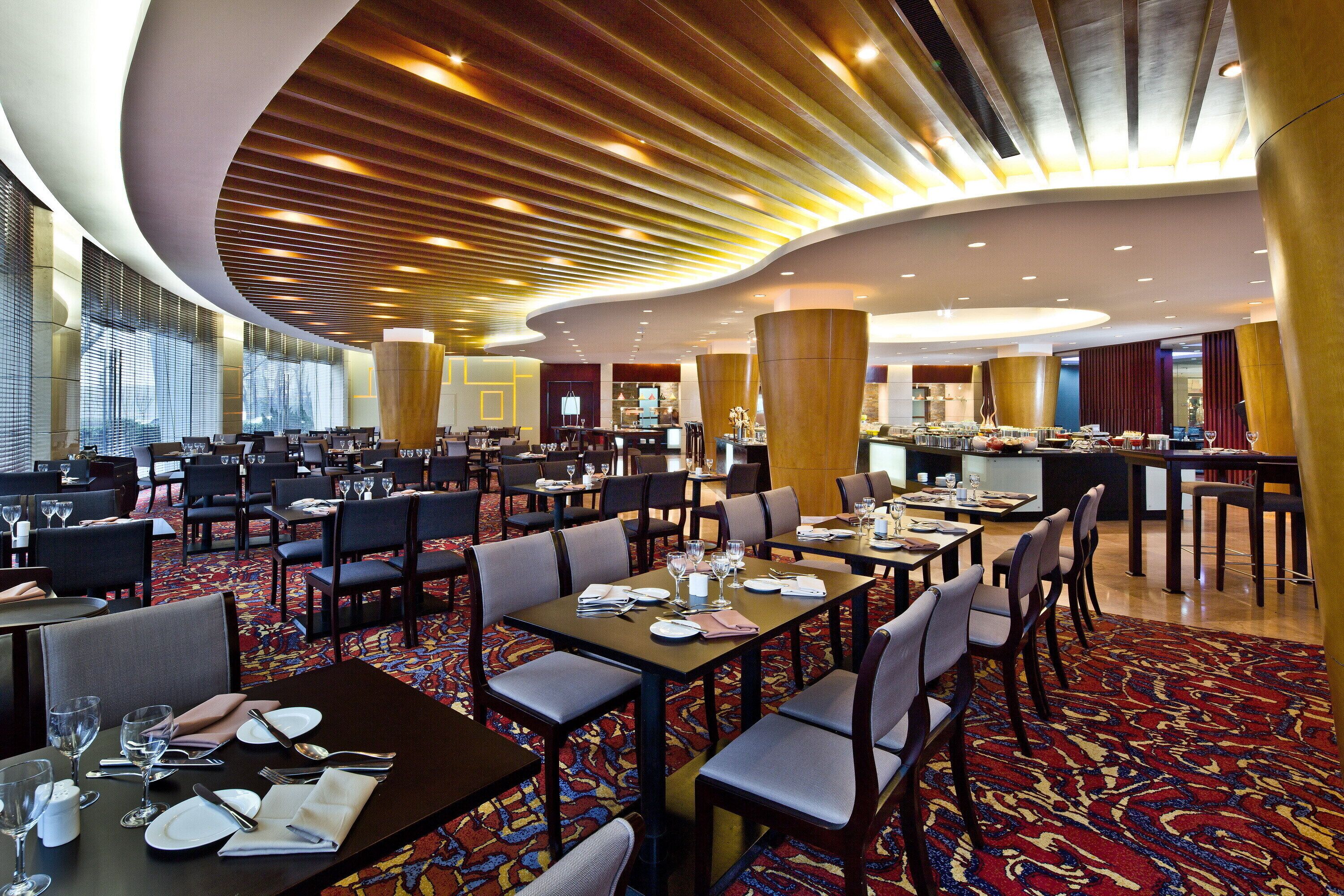 A spacious hotel dining area with multiple tables and chairs, a central bar, and a modern ceiling design.