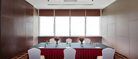 A conference room with a long table, white chairs, and red tablecloths. There are flower vases on the table and a window with blinds in the background.
