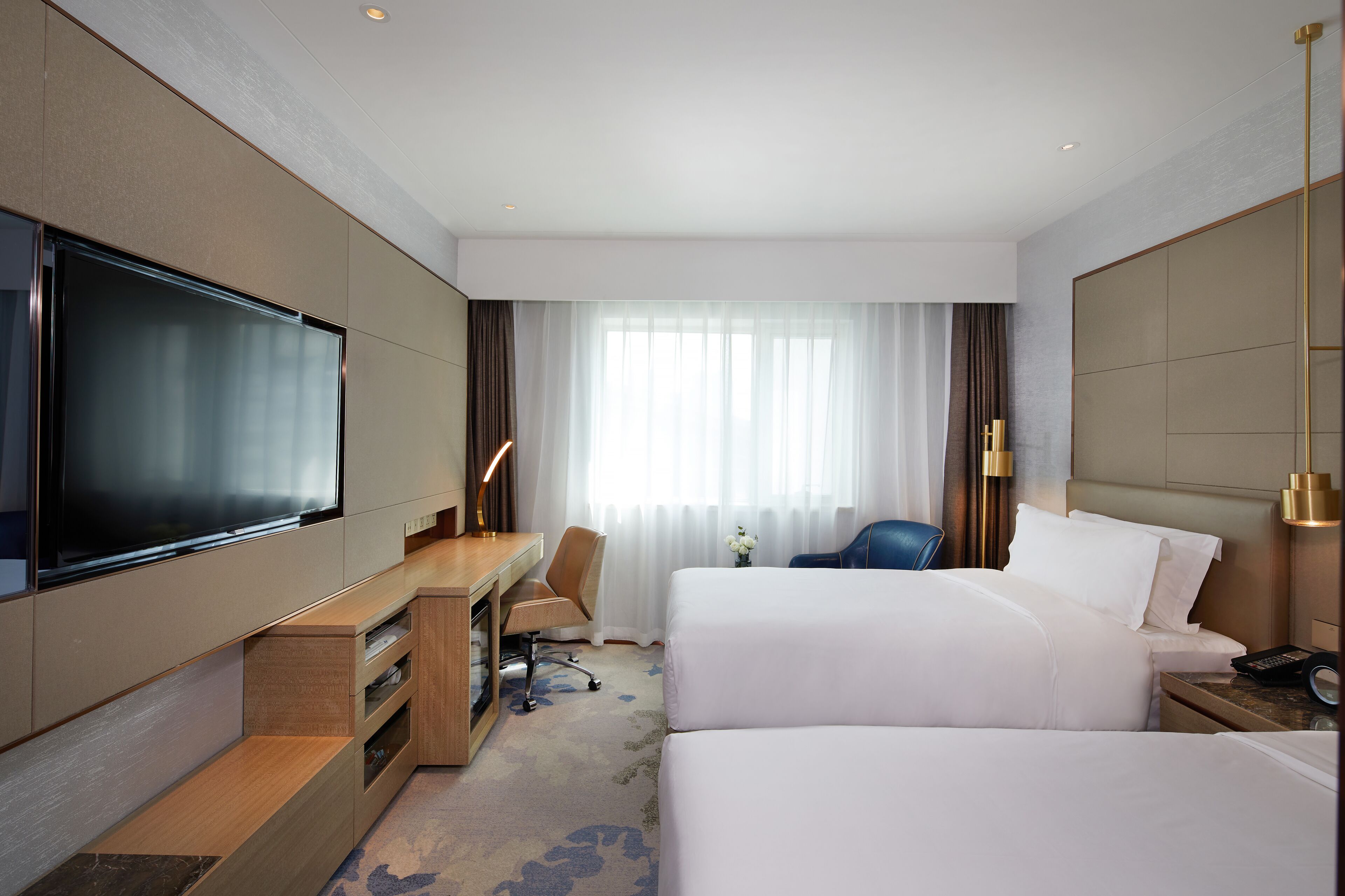 Habitación de hotel con televisión de pantalla plana, un escritorio con silla, dos camas, una mesita de noche con teléfono y un ventanal con cortinas.