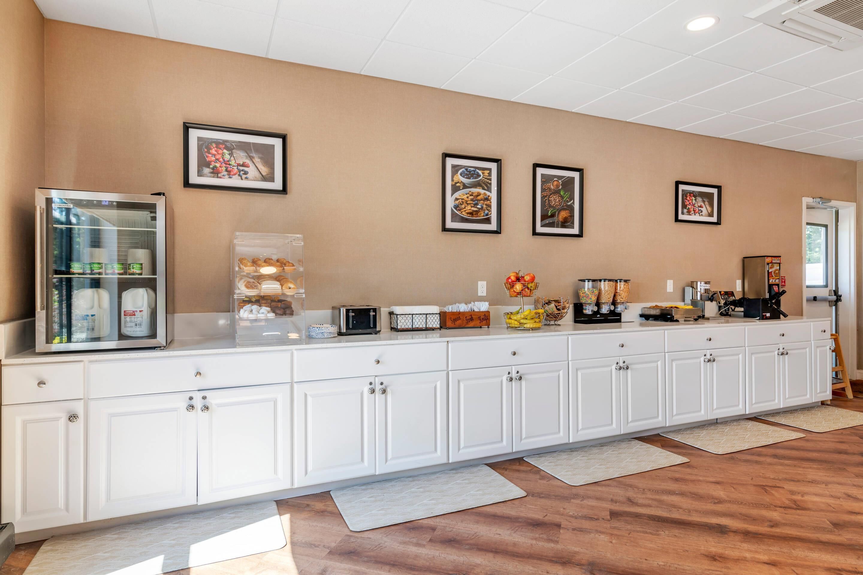 A long white counter with various food items and a refrigerator, framed pictures on the wall, and a wooden floor.
