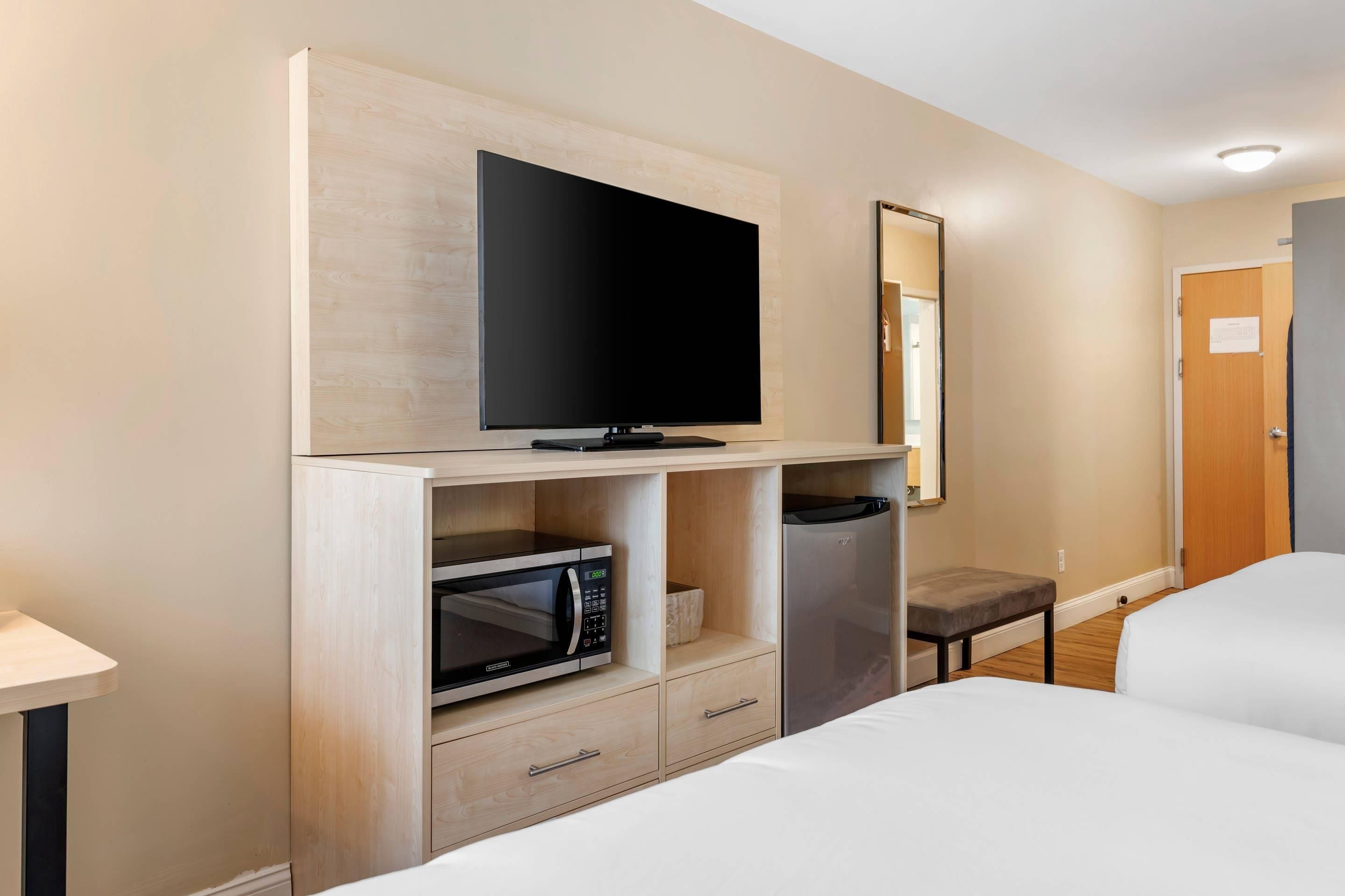 A hotel room with a flat-screen TV mounted on a wooden shelf, a microwave, a mini-fridge, a bedside table, and a large bed.