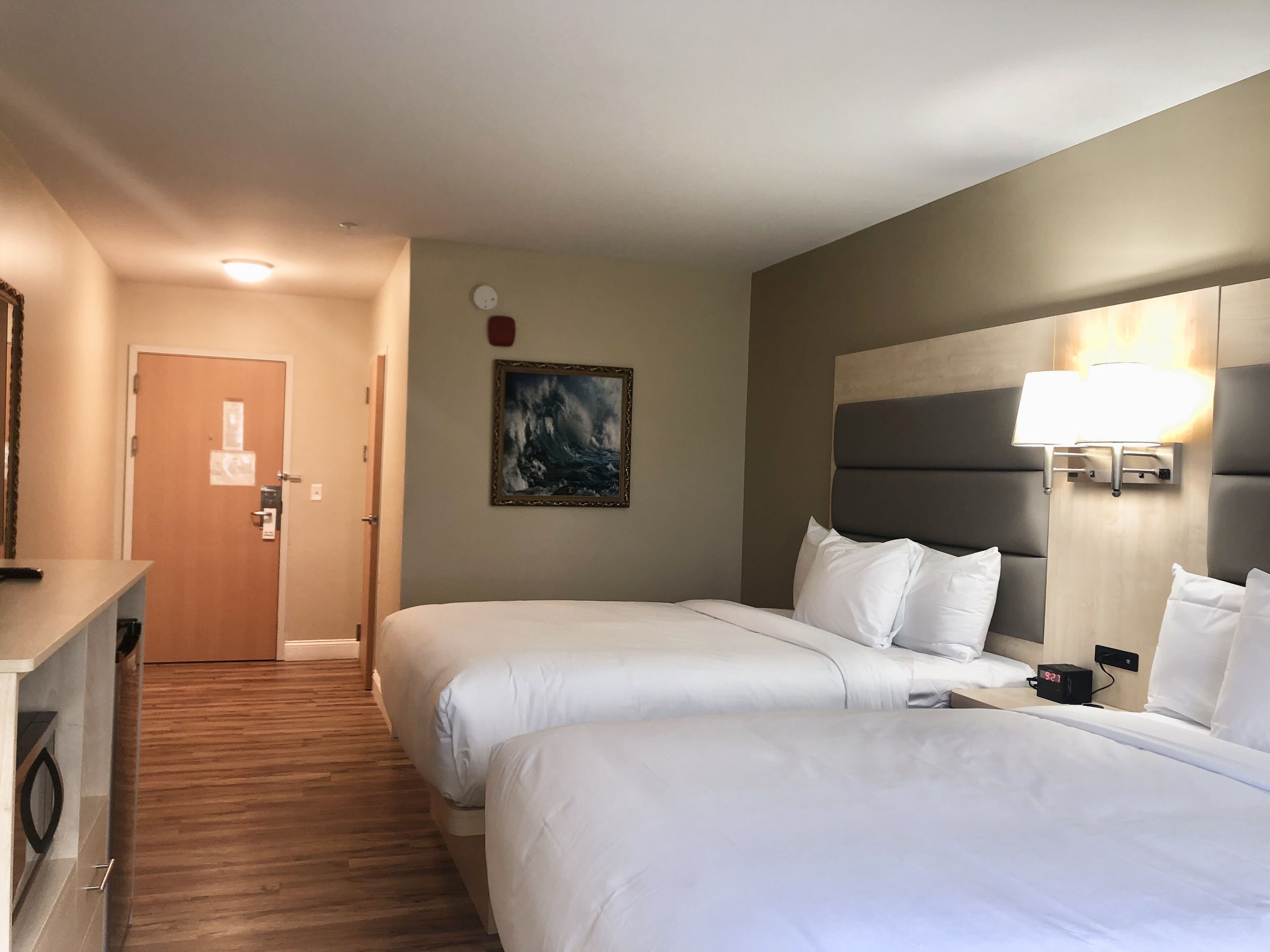 A hotel room with two beds, a painting on the wall, a clock, and a lamp on the nightstand.