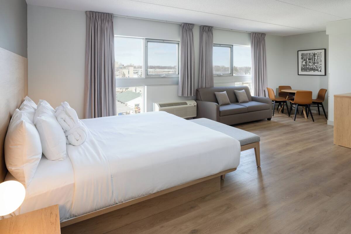 Ein Hotelzimmer mit einem großen Bett, einer Couch, einem Esstisch mit Stühlen und Blick auf die Stadt durch große Fenster.