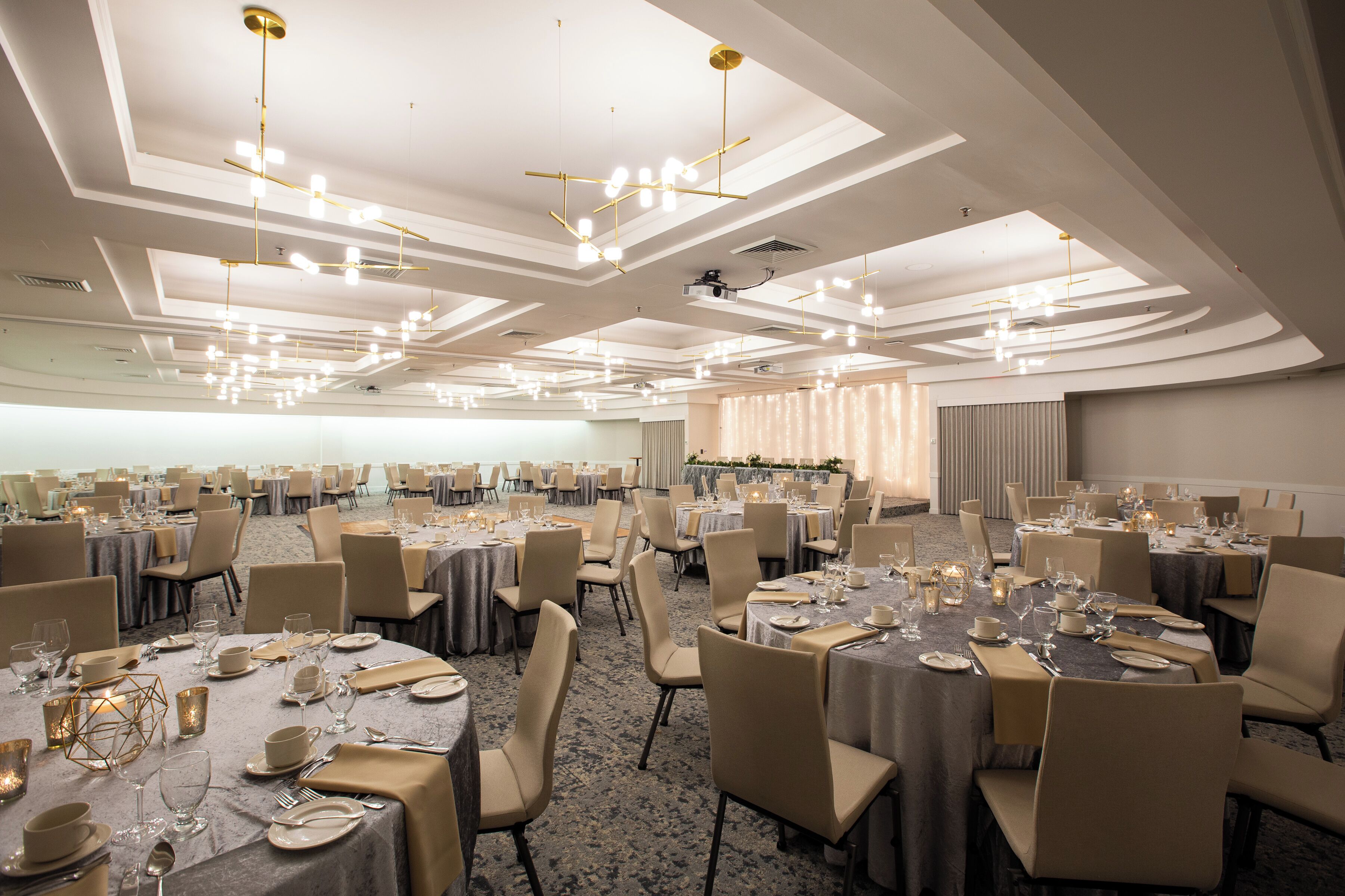 Une salle de banquet avec des tables rondes dressées pour le repas, agrémentée d’un lustre moderne et d’un plafond haut doté d’un éclairage encastré.