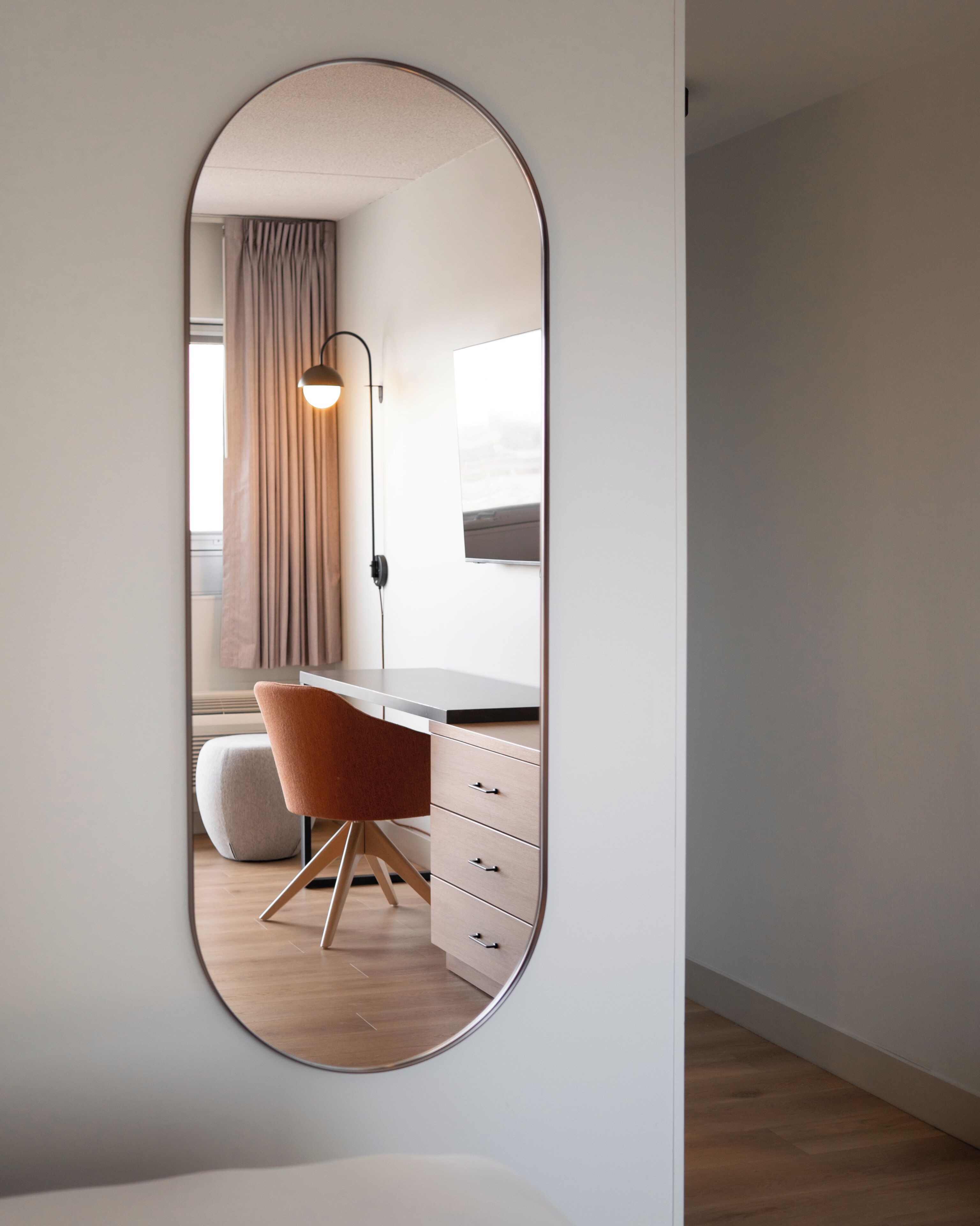 A room with a desk, chair, and a mirror reflecting a window with curtains.