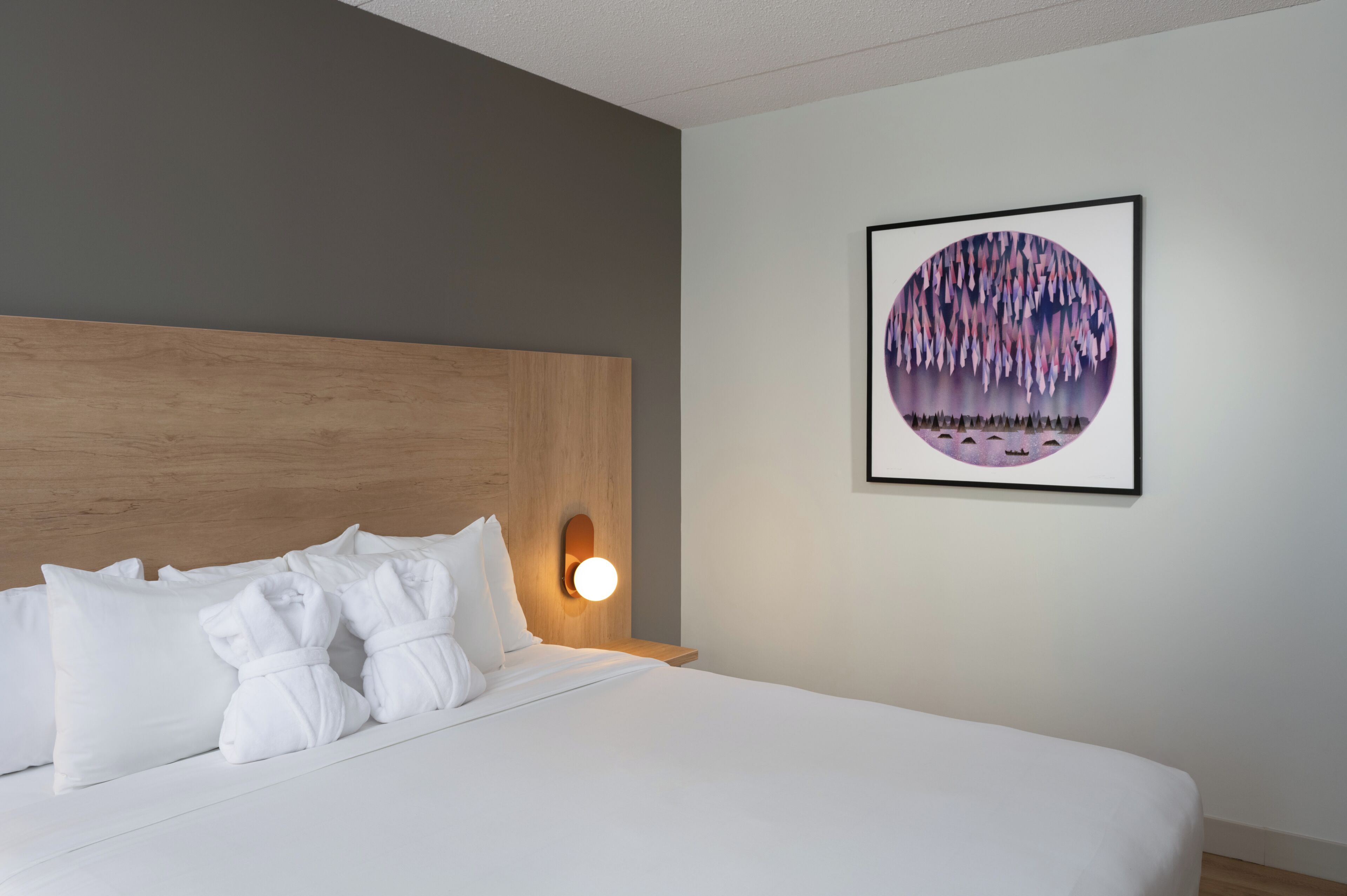 Una habitación de hotel con una cama, almohadas blancas, un cabecero, una lámpara fijada a la pared y una obra de arte enmarcada.