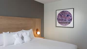 Una habitación de hotel con una cama, almohadas blancas, un cabecero, una lámpara fijada a la pared y una obra de arte enmarcada.