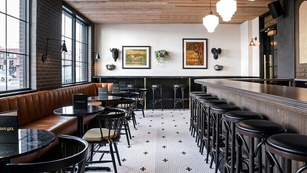 Un área de bar con asientos de cuero, mesas redondas y taburetes de bar. Hay cuadros en la pared y una barra con un letrero encima.