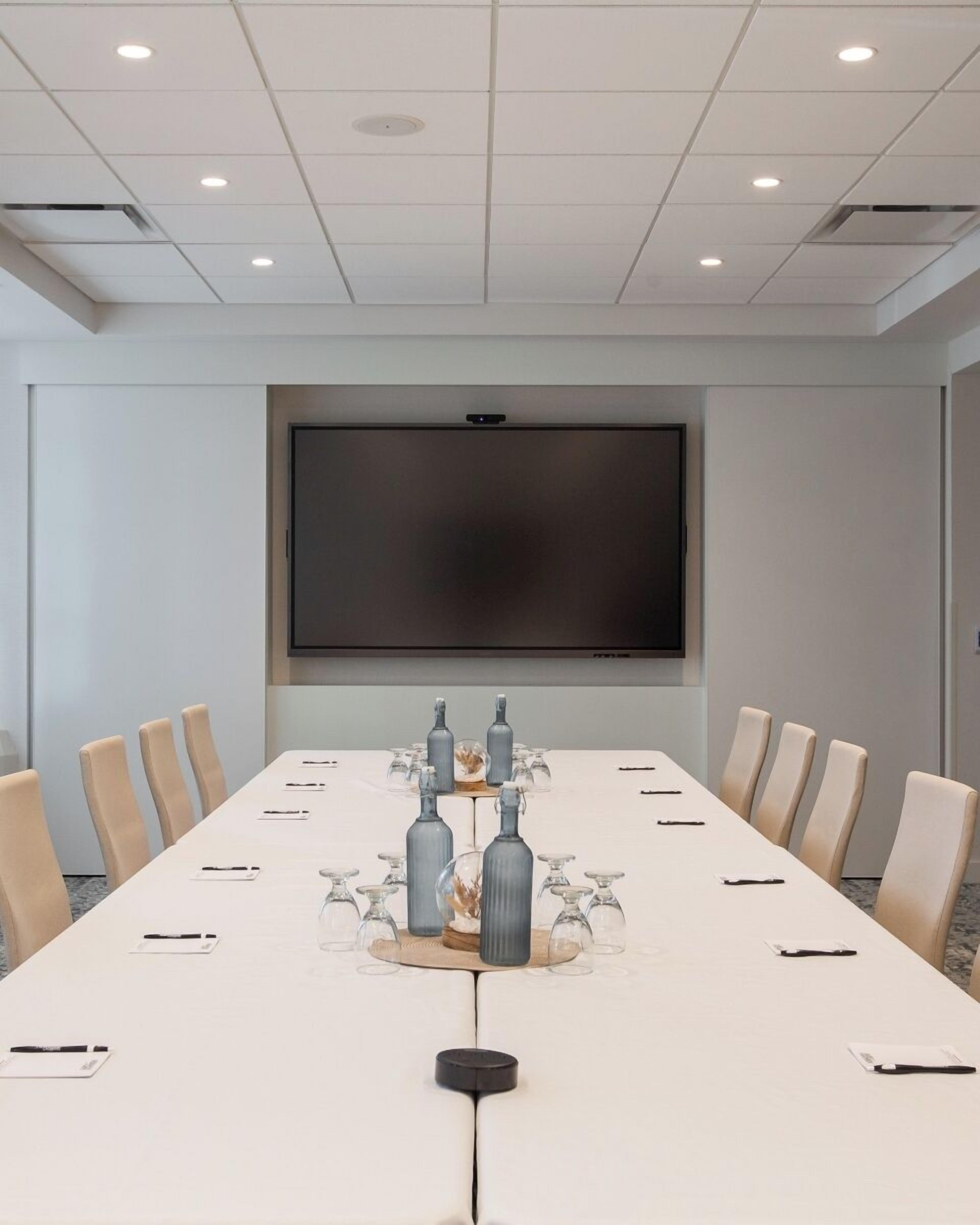 A conference room with a long table, beige chairs, a screen, and decorative bottles.