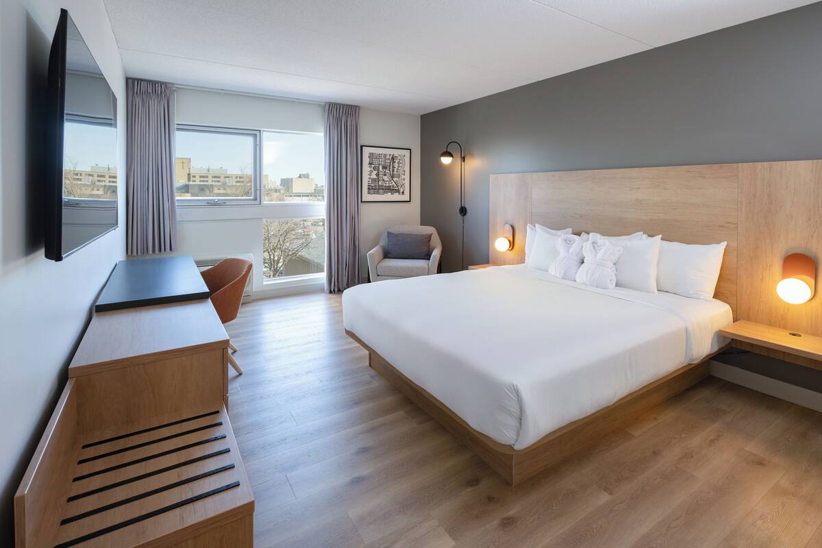 A hotel room with a large bed, a desk, a chair, a TV, and a city view.