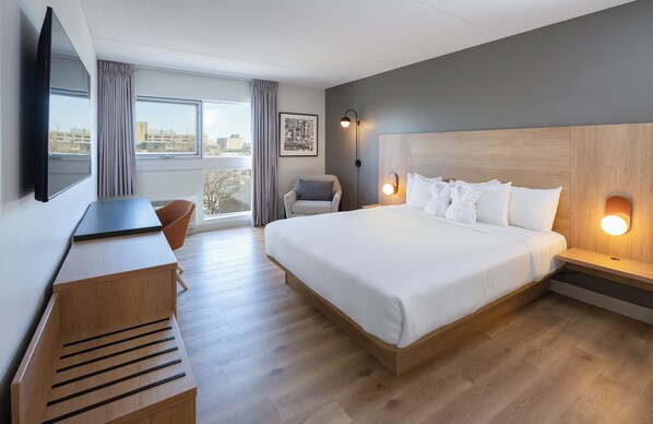 A hotel room with a large bed, a desk, a chair, a TV, and a city view. - Norwood Hotel (Winnipeg)