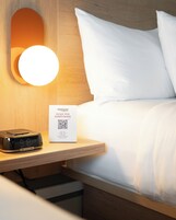 Habitación de hotel con mesita de noche, reloj digital, cartel con código QR y cama con ropa blanca.