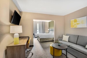 A hotel room with a bed, a sofa, a desk with a lamp, a TV, and a painting on the wall.
