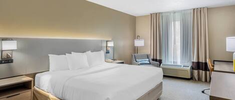 A hotel room with a large bed, bedside tables, a desk, a chair, a window with curtains, and an air conditioning unit.