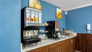 Una estación de desayuno autoservicio con dispensadores de jugo, una máquina de café y un refrigerador con alimentos.