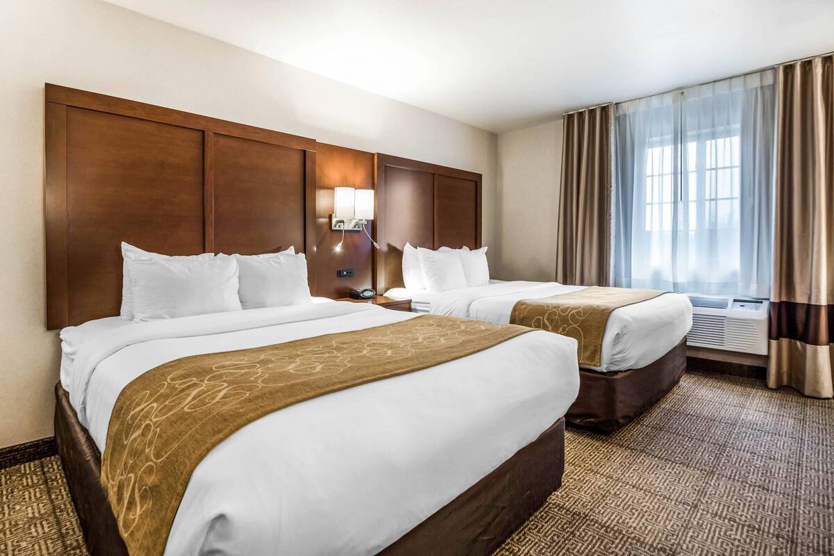 A hotel room with two beds, a large wooden headboard, a wall-mounted lamp, and a large window with curtains.