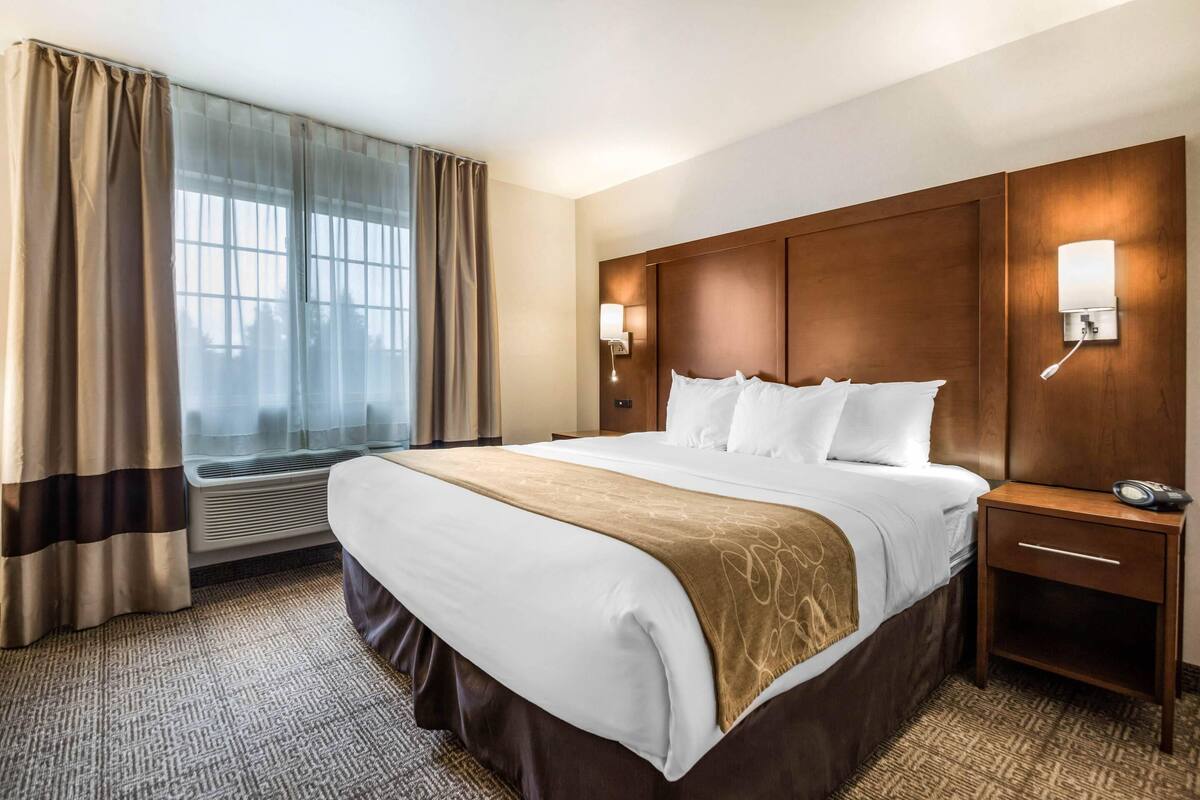 A hotel room with a large bed, a wooden headboard, two bedside lamps, a nightstand, and a window with curtains.