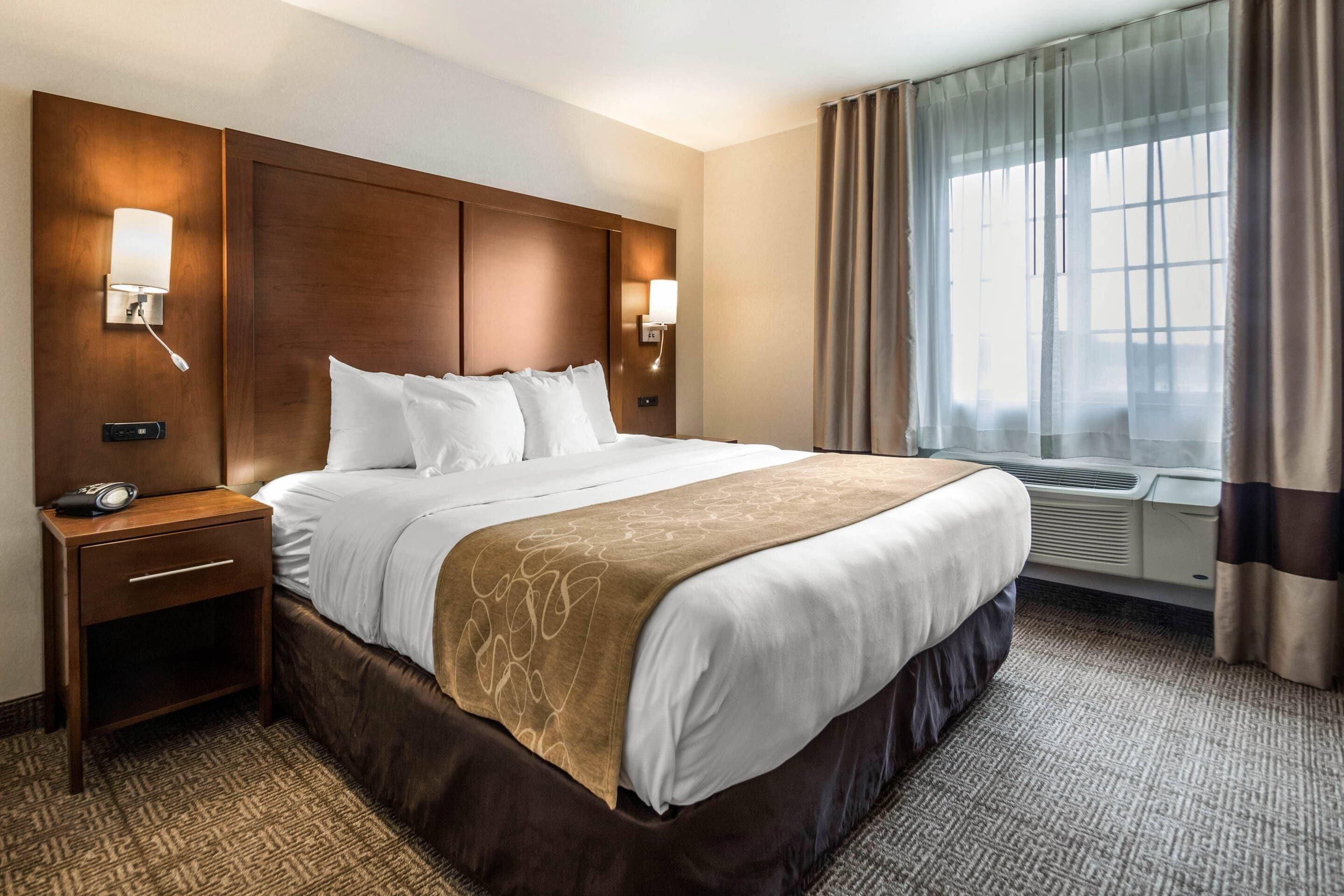 A hotel room with a large bed, bedside lamps, an air conditioning unit, a nightstand, and a window with curtains.