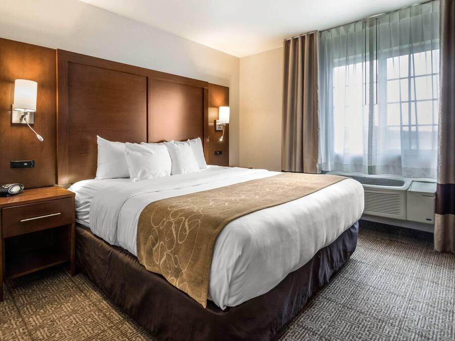 A hotel room with a large bed, bedside lamps, an air conditioning unit, a nightstand, and a window with curtains.