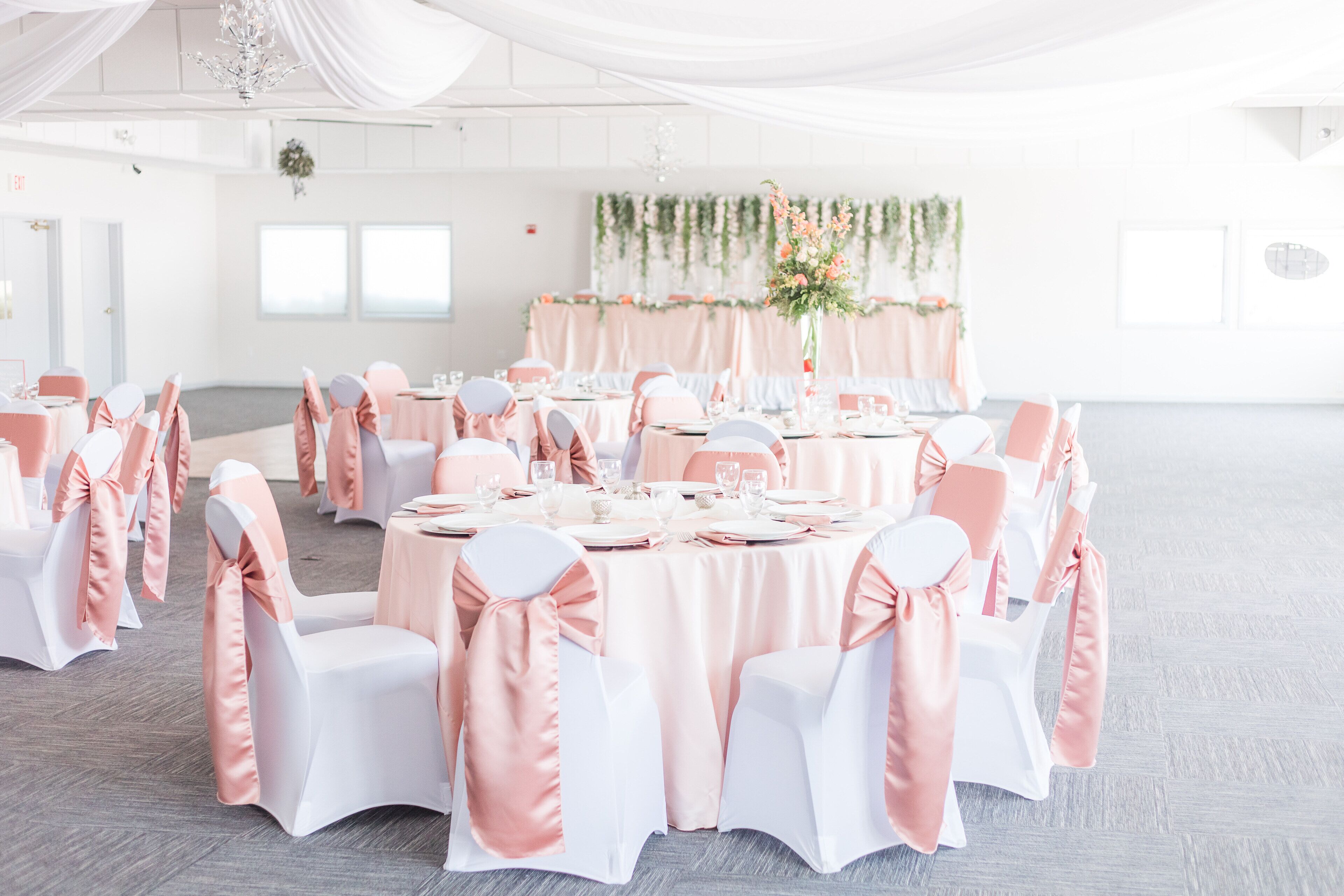 A banquet hall with round tables, white and pink chairs, and a floral centerpiece.