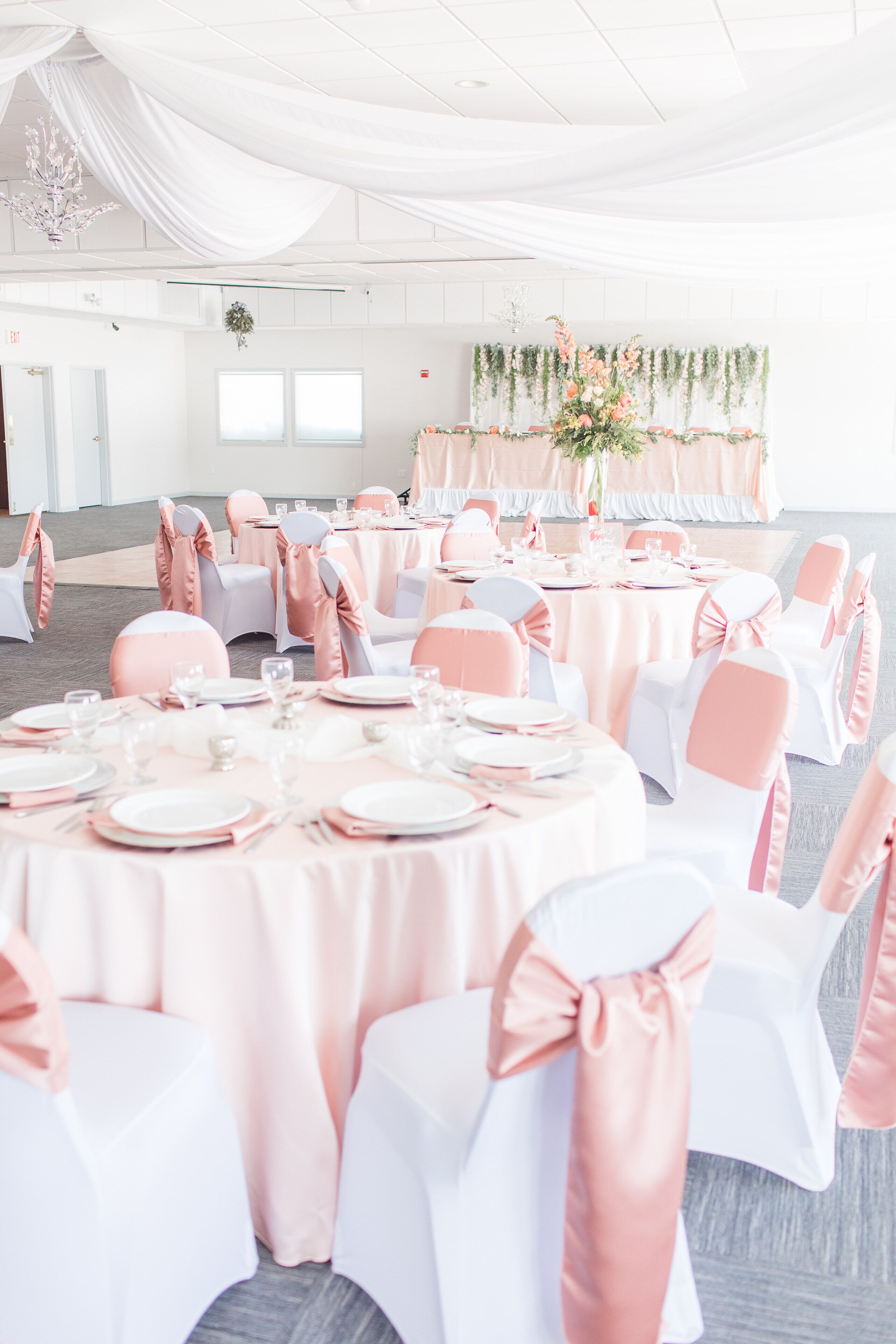 A banquet hall with round tables, white and pink chairs, and floral arrangements.