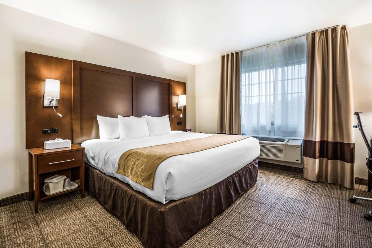A hotel room with a large bed, bedside tables, a nightstand, a window with curtains, and an air conditioning unit.