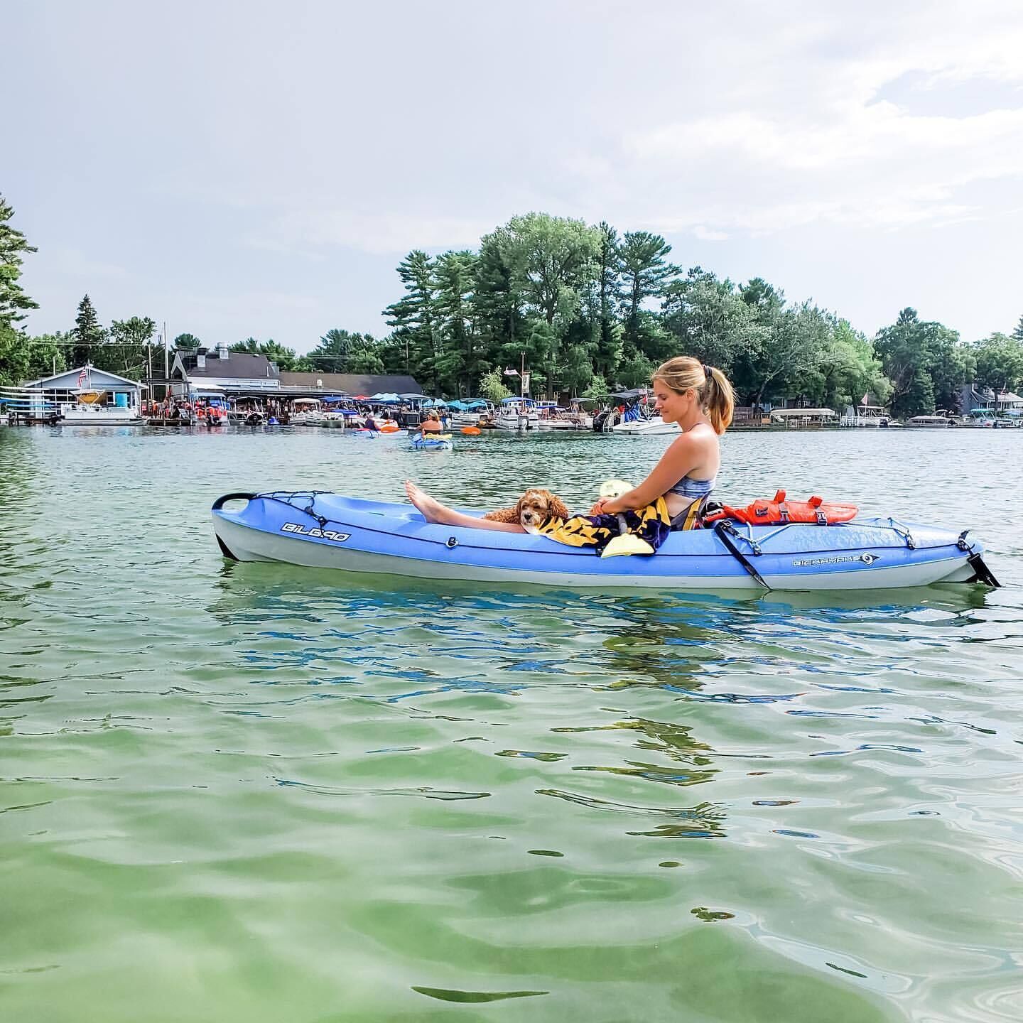 A person in a blue kayak on calm water with a dog, surrounded by trees and a dock with people and boats in the background.