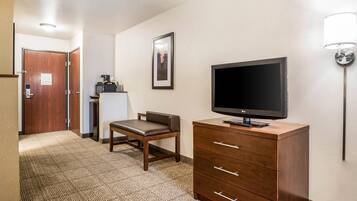 A hotel room with a TV on a wooden cabinet, a desk with a chair, a framed picture on the wall, and a lamp.