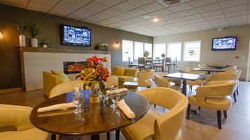 A hotel dining area with multiple tables and chairs, a fireplace, and two televisions mounted on the walls.