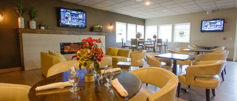 A hotel dining area with multiple tables and chairs, a fireplace, and two televisions mounted on the walls.
