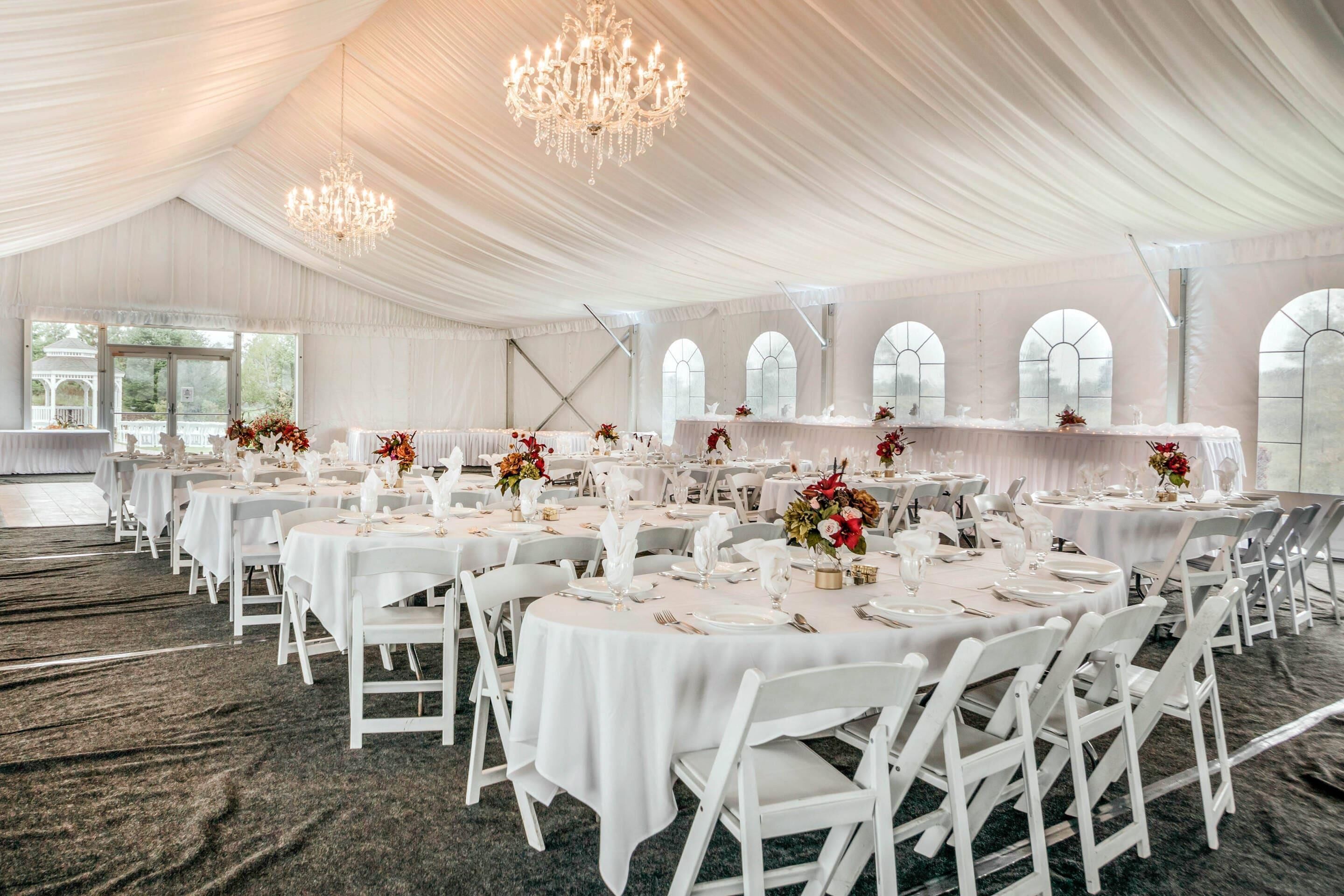 Une salle de banquet avec des tables rondes, des chaises blanches, des lustres et de grandes fenêtres.