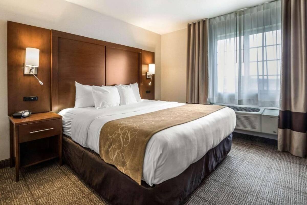 A hotel room with a large bed, two bedside lamps, a nightstand, a window with curtains, and an air conditioning unit.