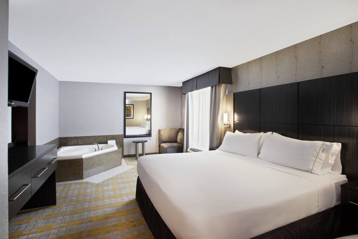 Ein Hotelzimmer mit einem großen Bett, einem separaten Badezimmer mit Badewanne, einem Flachbildfernseher, einem Sessel und einem Spiegel.