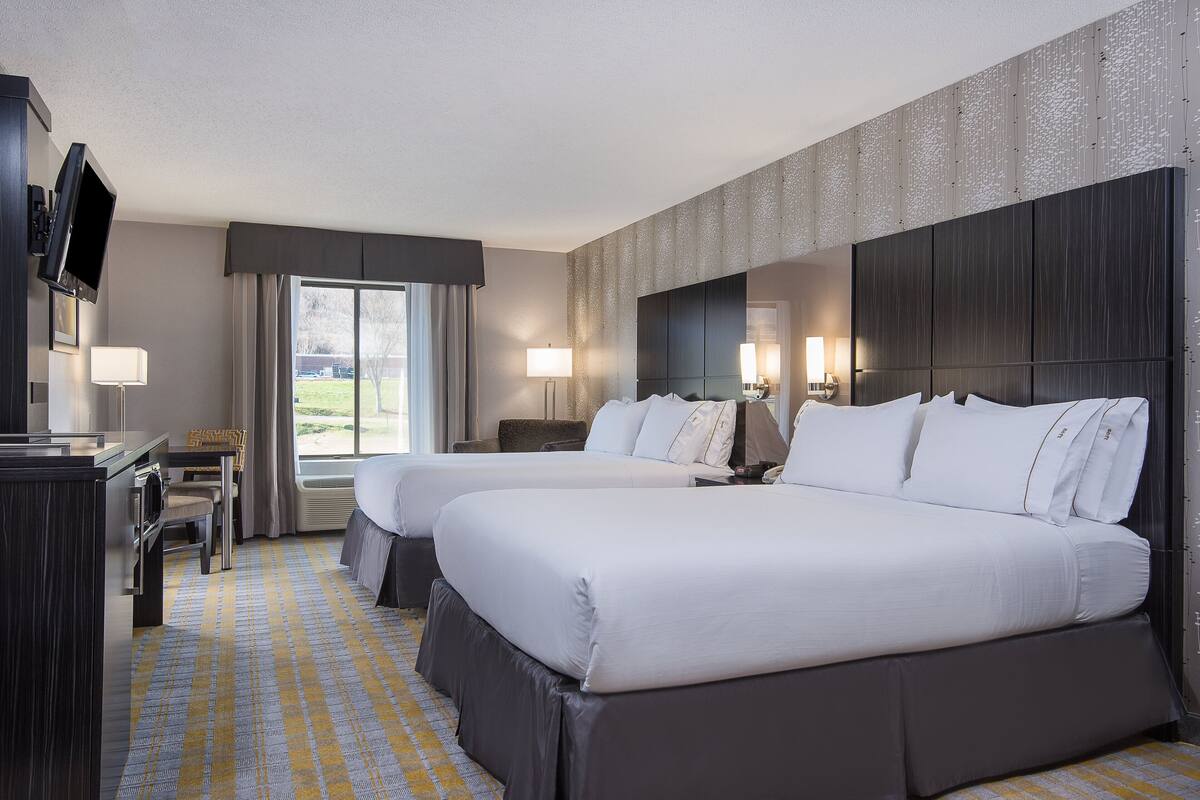 Ein Hotelzimmer mit zwei Betten, einem Schreibtisch, einem Fernseher und einem großen Fenster mit Vorhängen.