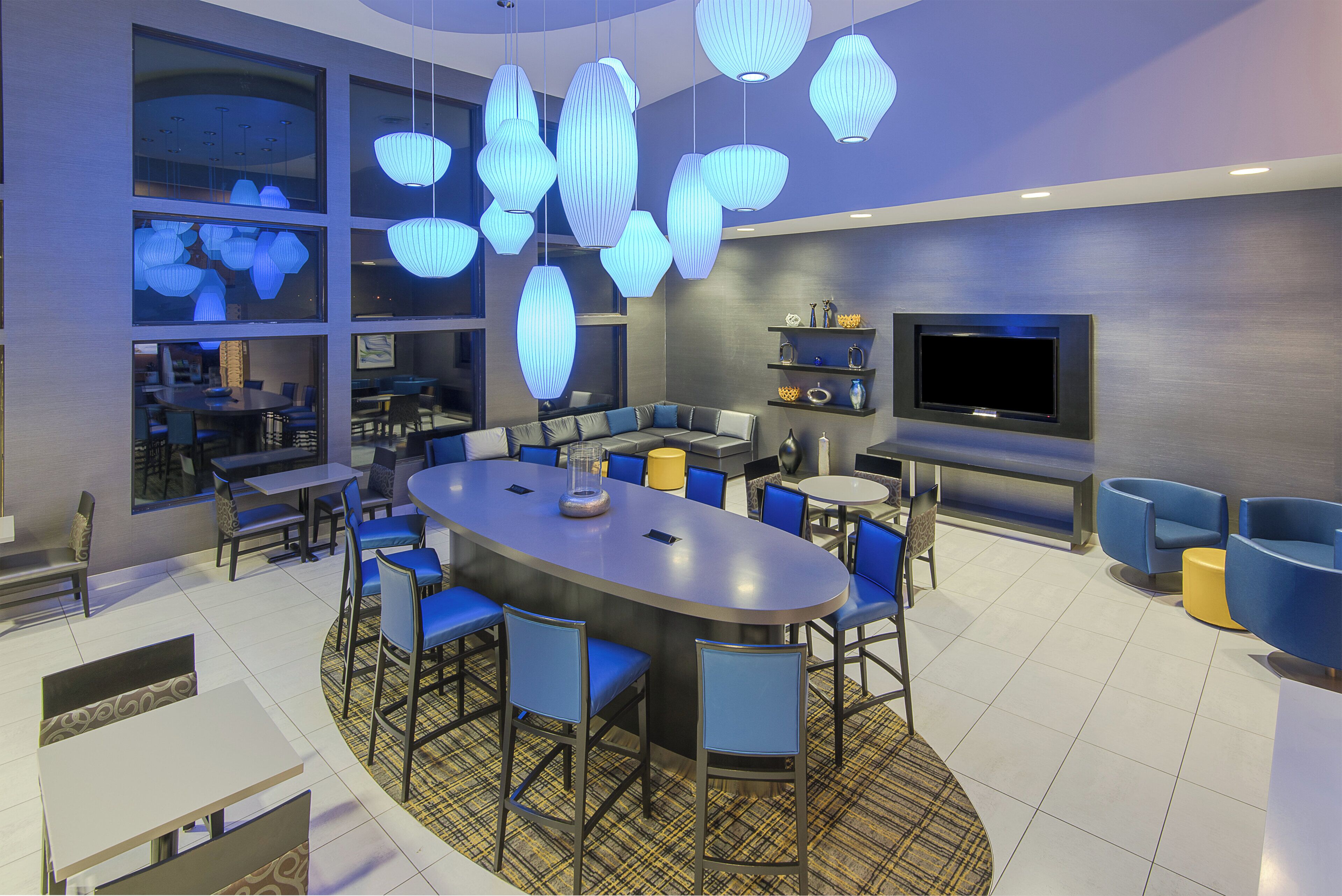 A modern interior with a central round table surrounded by chairs, a TV mounted on the wall, and a seating area with sofas and chairs.