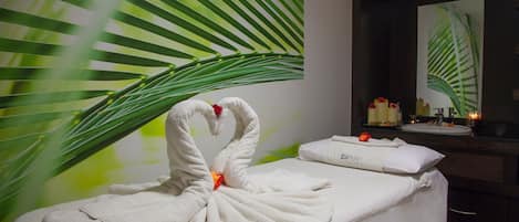 A spa room with a massage table, a towel swan decoration, a large green leaf wall, a mirror, and a sink with candles.