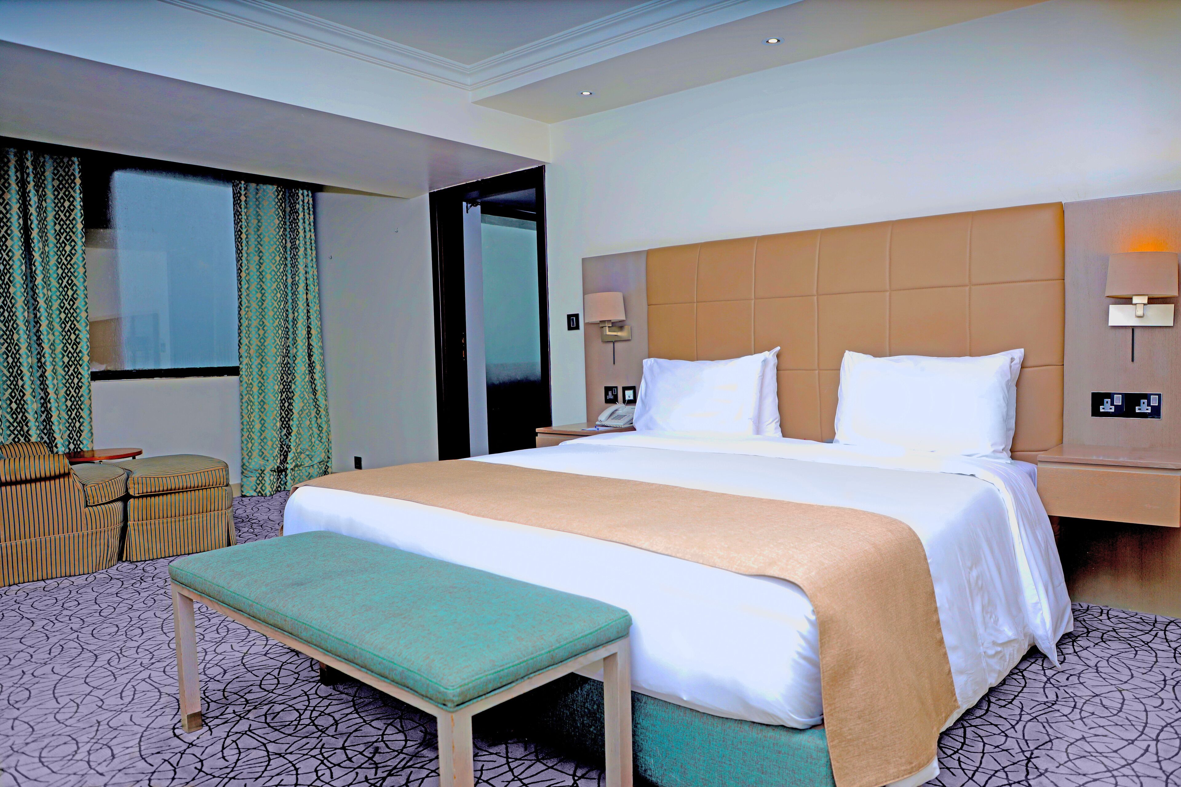 Una habitación de hotel con una cama grande, un banco, una mesita y una ventana con cortinas.