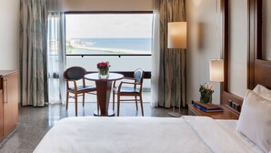 A hotel room with a large bed, a desk with a chair, a small table with a vase of flowers, and a view of the ocean. - Eko Hotel Main Building (Lagos)