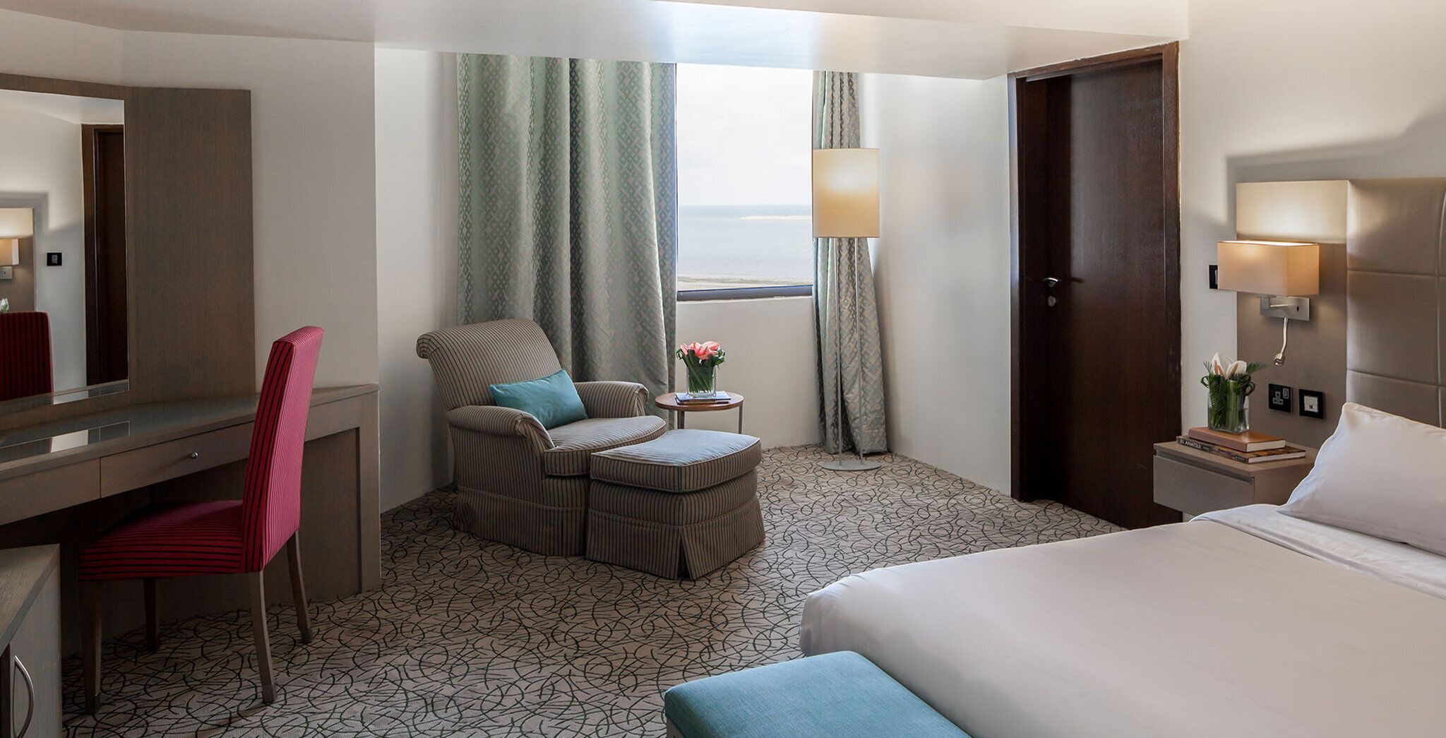 A hotel room with a bed, a desk with a chair, a sofa with a pillow, a small table with a lamp, and a view of the sea through a window.