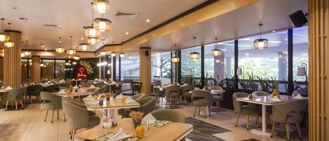 A restaurant with multiple tables and chairs, large windows, and hanging pendant lights.